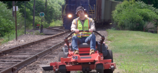 a man on a riding mower near railroad tracks with a train in the background